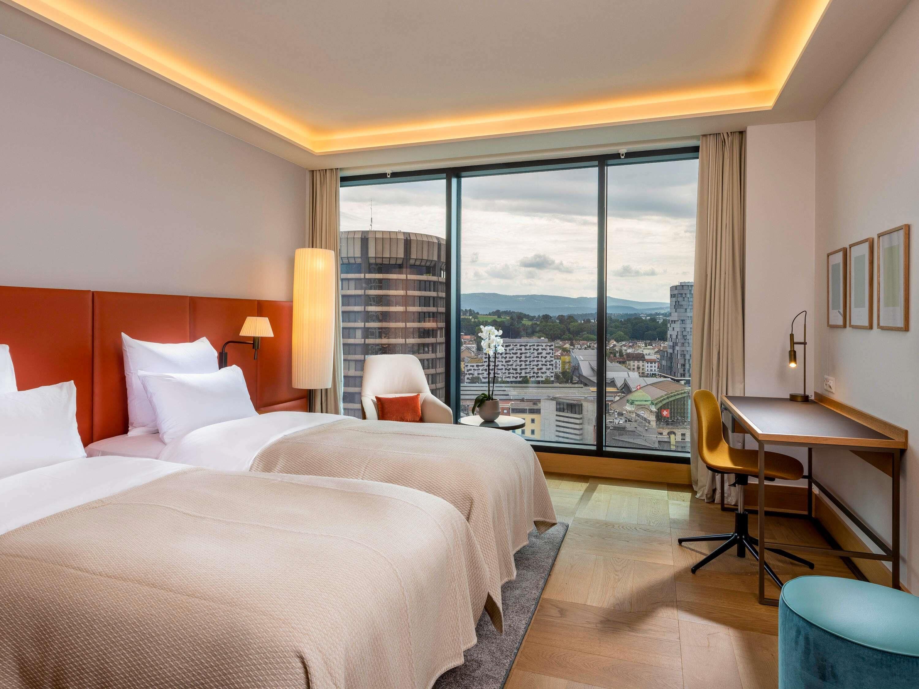 A hotel room with two beds, a desk, a chair, a lamp, and a large window offering a city view.
