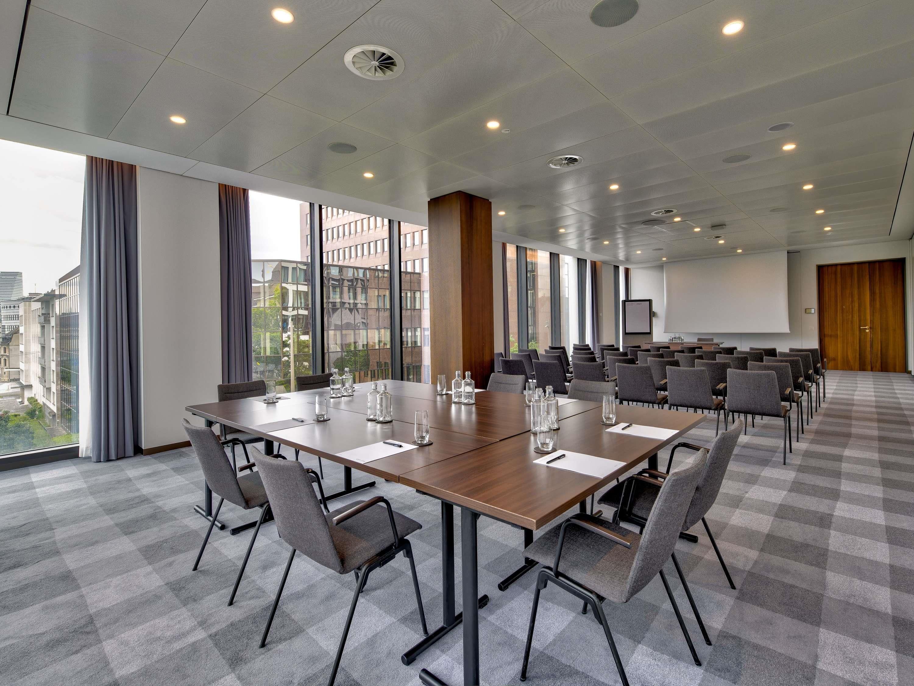 Un salón de conferencias con una gran mesa, sillas, una pantalla de proyector y ventanas amplias con cortinas.