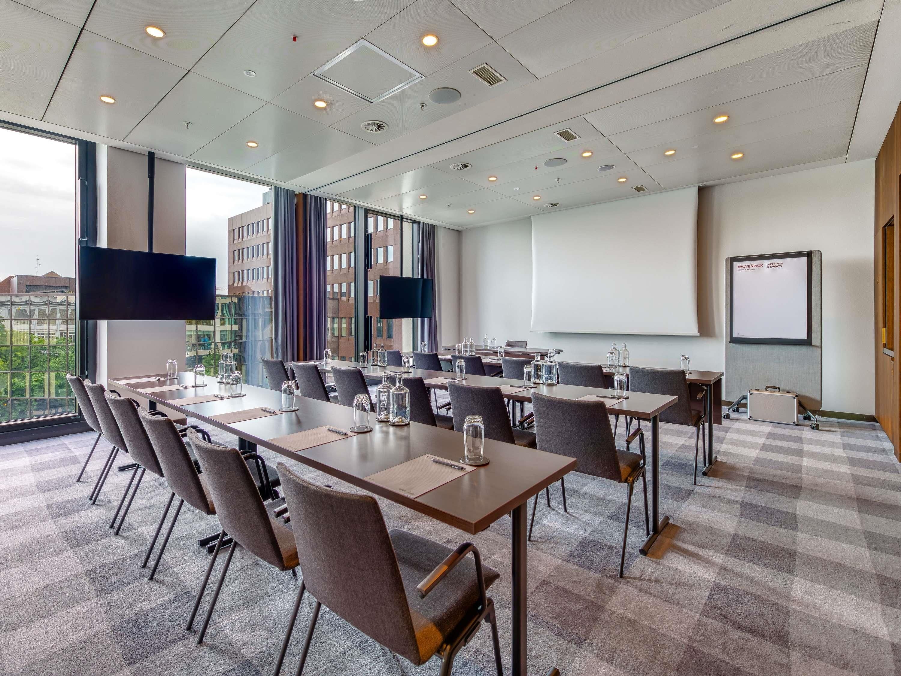 A conference room with a large table, chairs, a projector screen, and a view of buildings through large windows.