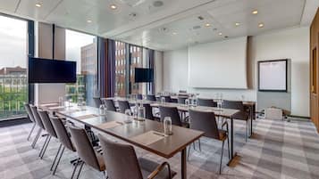 A conference room with a large table, chairs, a projector screen, and a view of buildings through large windows.