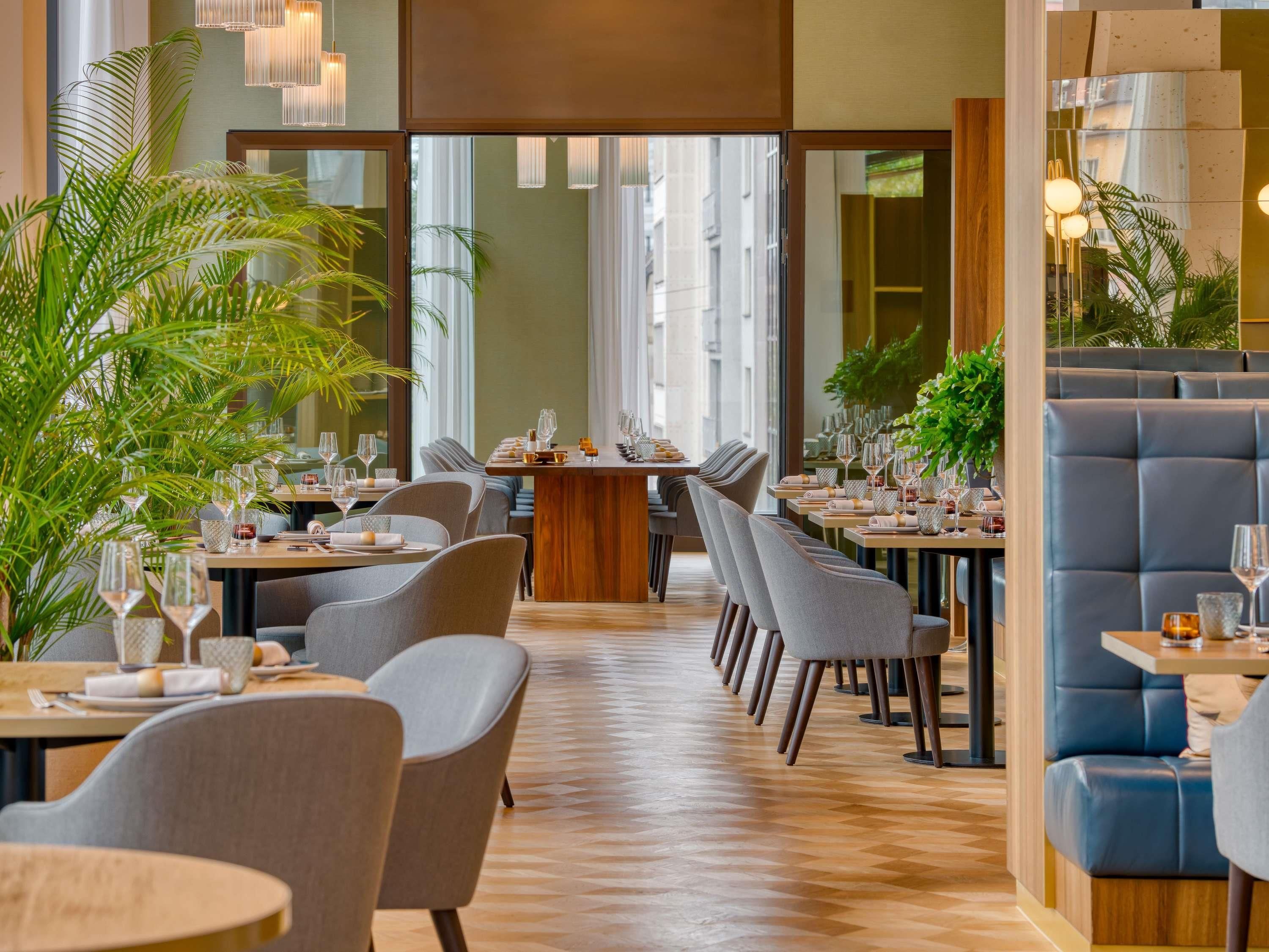 A modern restaurant interior with wooden flooring, grey upholstered chairs, and tables set for dining.