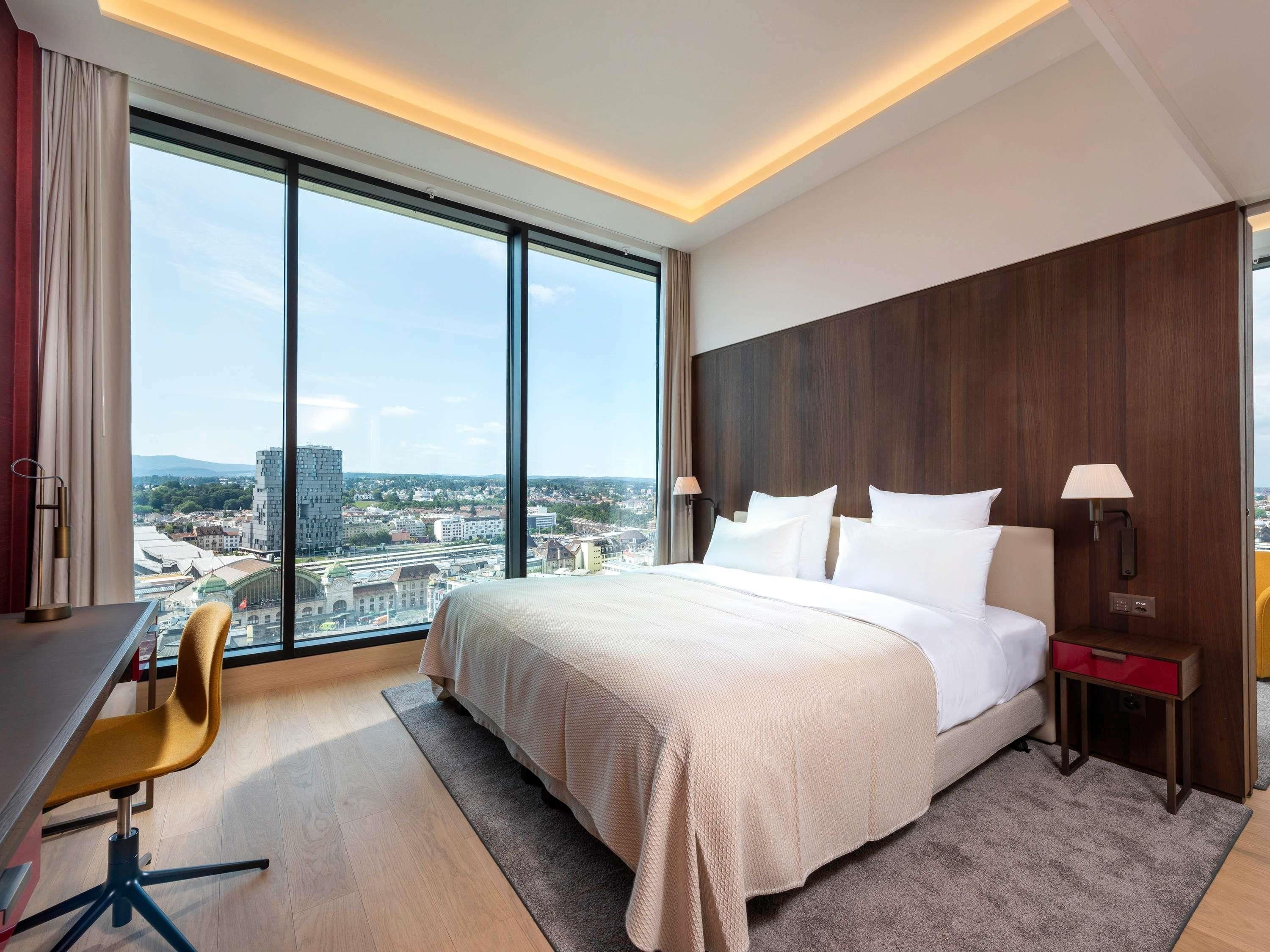Ein modernes Hotelzimmer mit einem großen Bett, einem Schreibtisch und einem Stuhl. Es bietet einen Blick auf die Stadt durch große Fenster und ein Holz-Kopfteil.