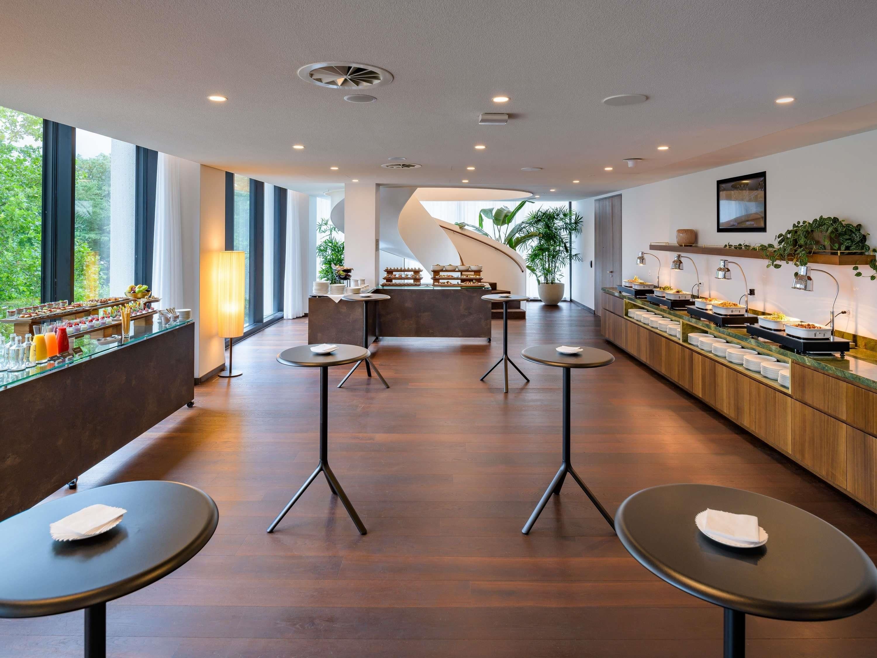 A modern hotel lobby with a central bar area, round tables, and a view of greenery outside.