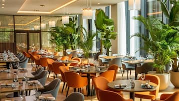 Un restaurante con varias mesas y sillas, plantas en macetas grandes y lámparas colgantes.