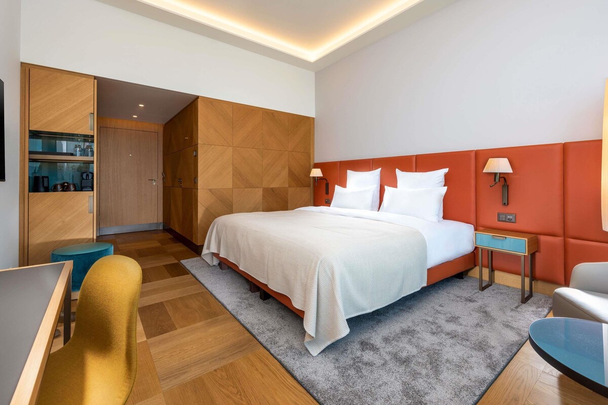 A modern hotel room with a large bed, a desk, a chair, and a wardrobe.