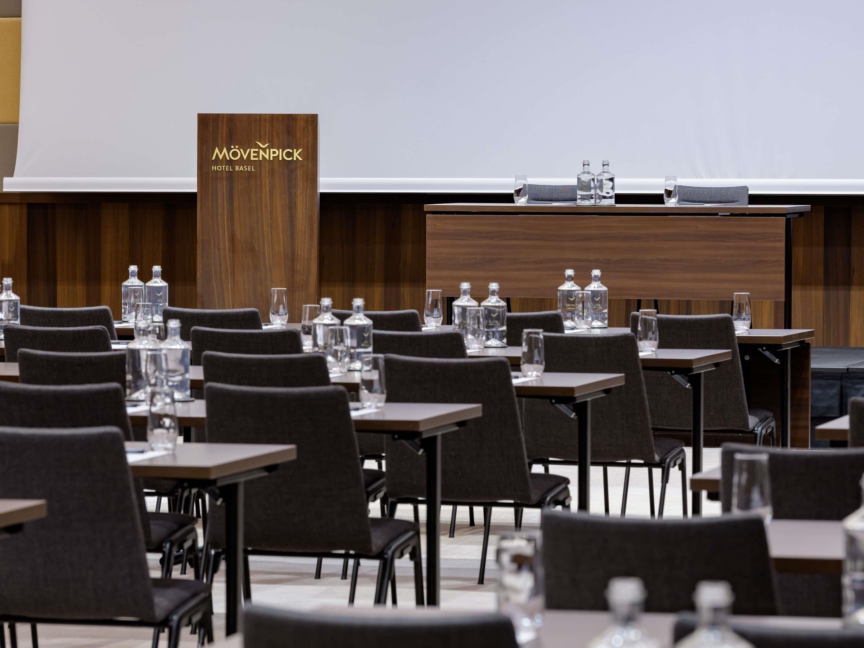 Un salón de conferencias con mesas y sillas, un atril con el nombre del hotel, y botellas de agua sobre las mesas.