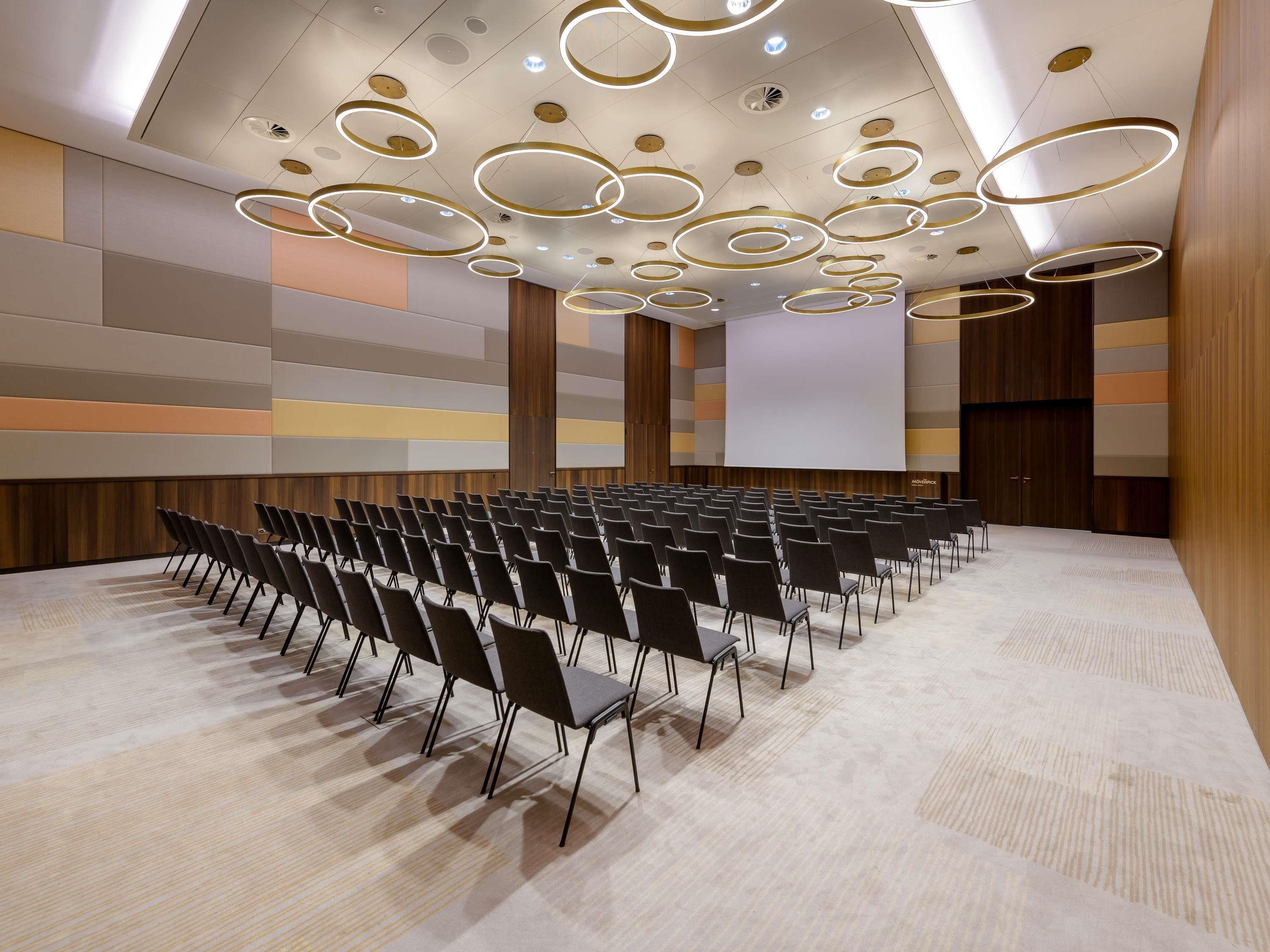 Un salón de conferencias con filas de sillas, una pantalla grande y luces circulares en el techo.