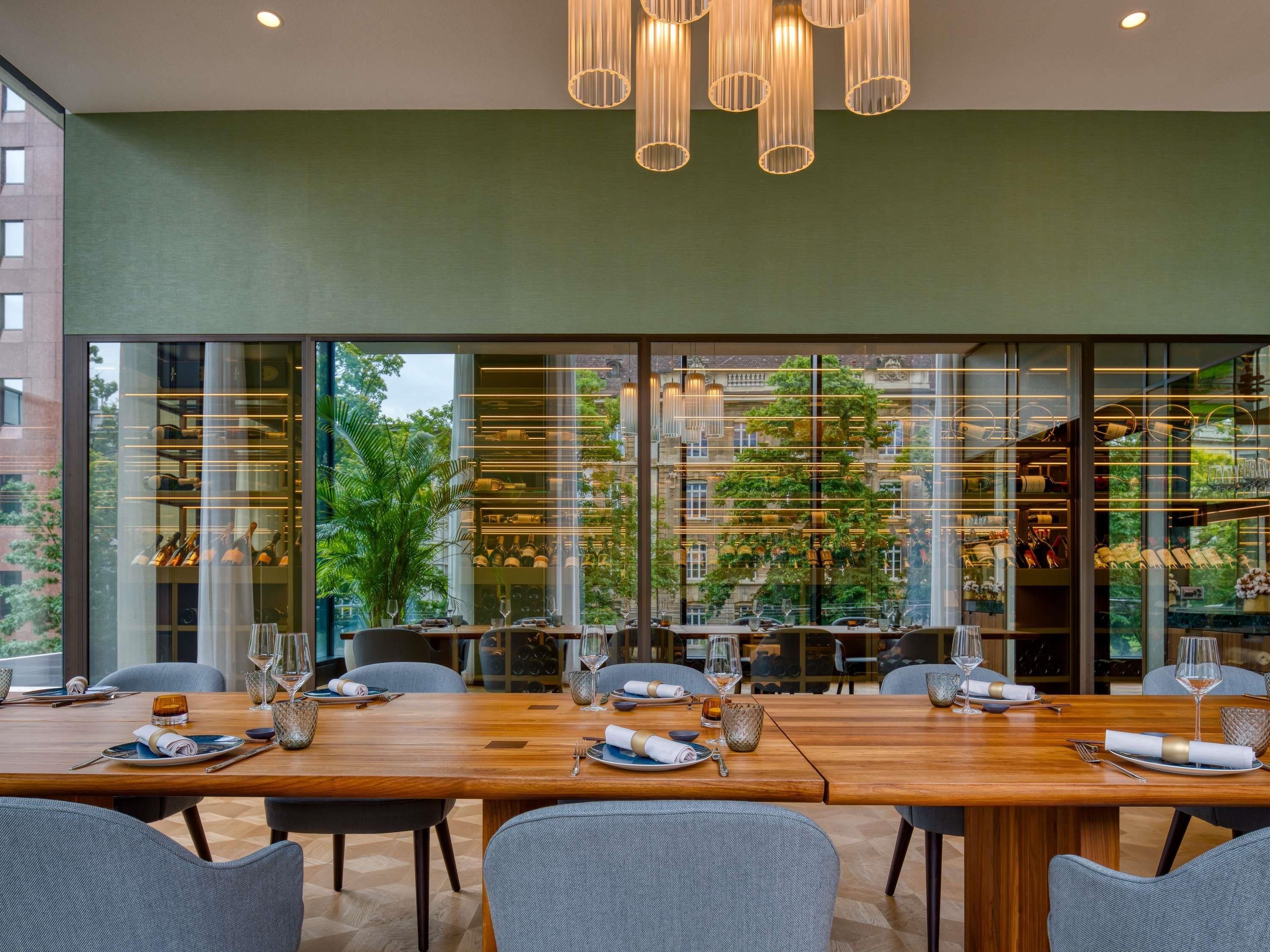 A modern dining area with a long wooden table, grey upholstered chairs, and a glass partition with shelves of bottles and glasses.