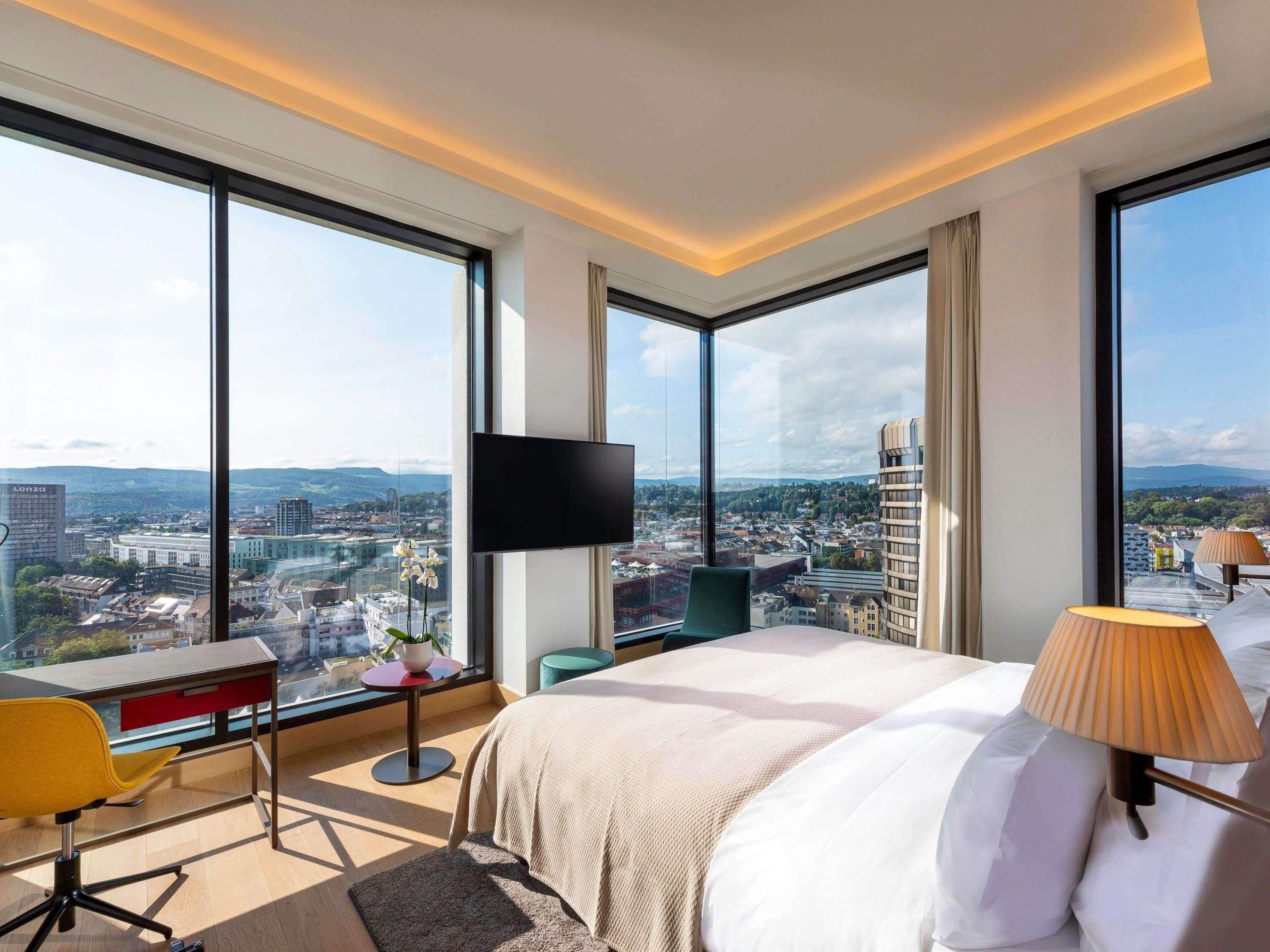 A hotel room with a large bed, a desk, a chair, a TV, and a view of the city.