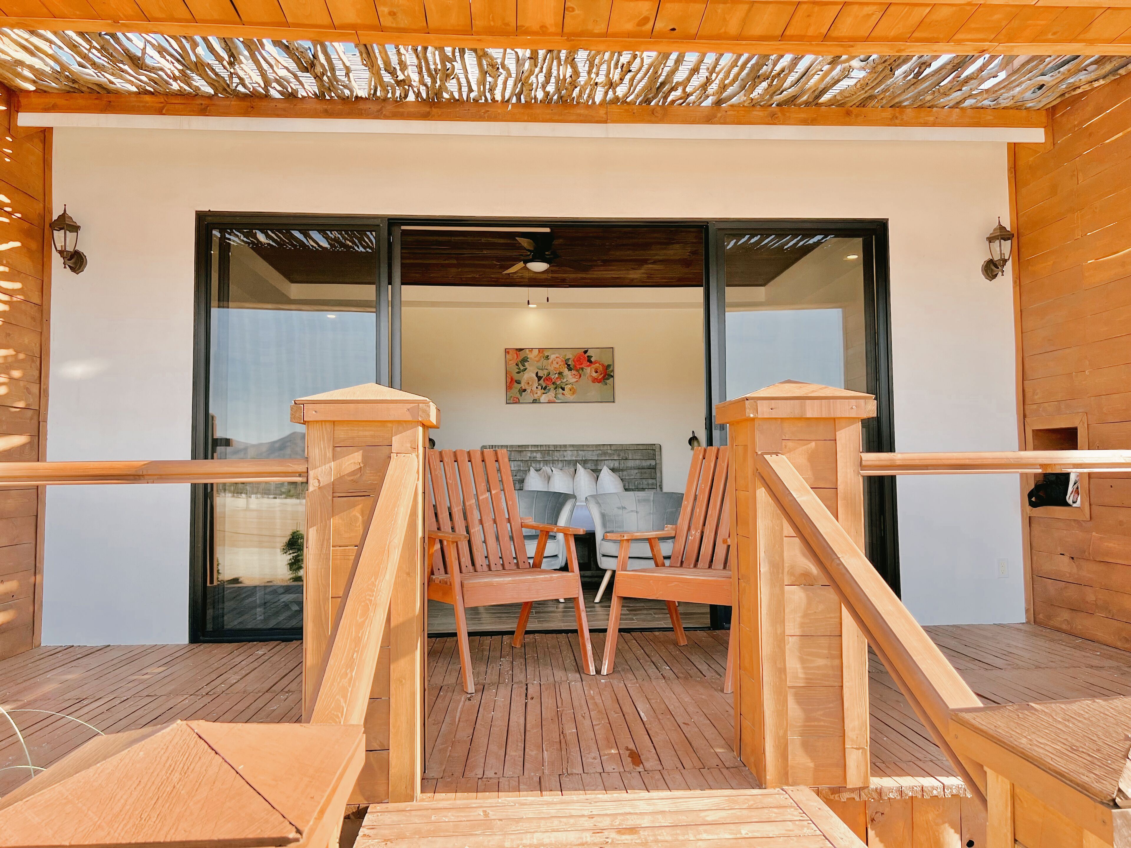 Una terraza de madera con un ventanal de vidrio que da a una habitación con una cama y un cuadro.