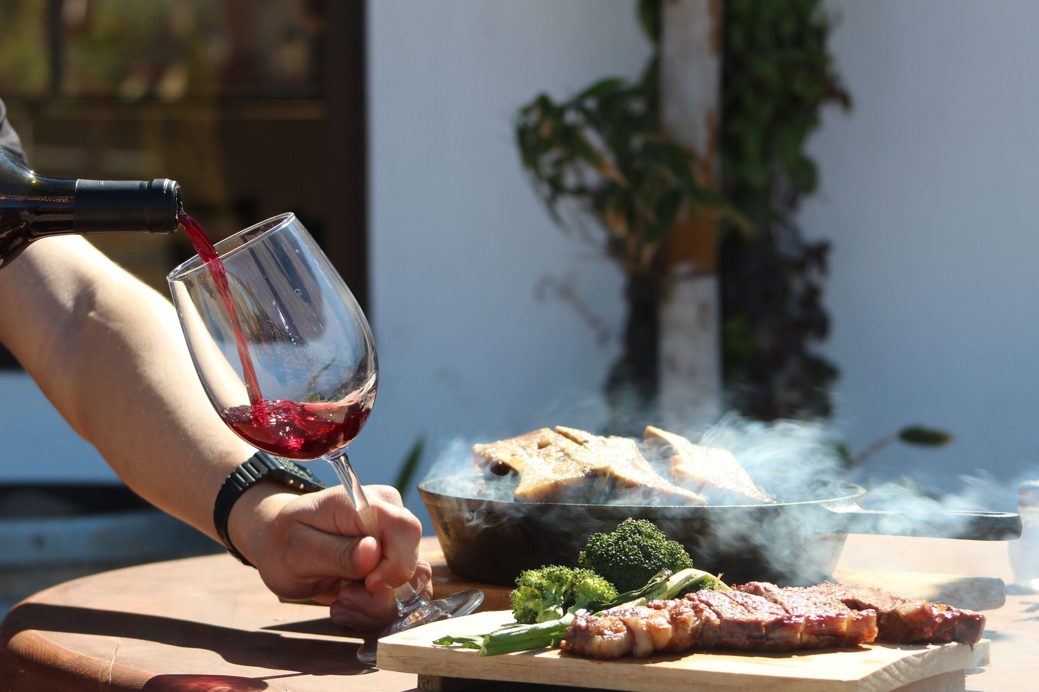 A person pouring red wine into a glass, with a grill cooking food in the background.