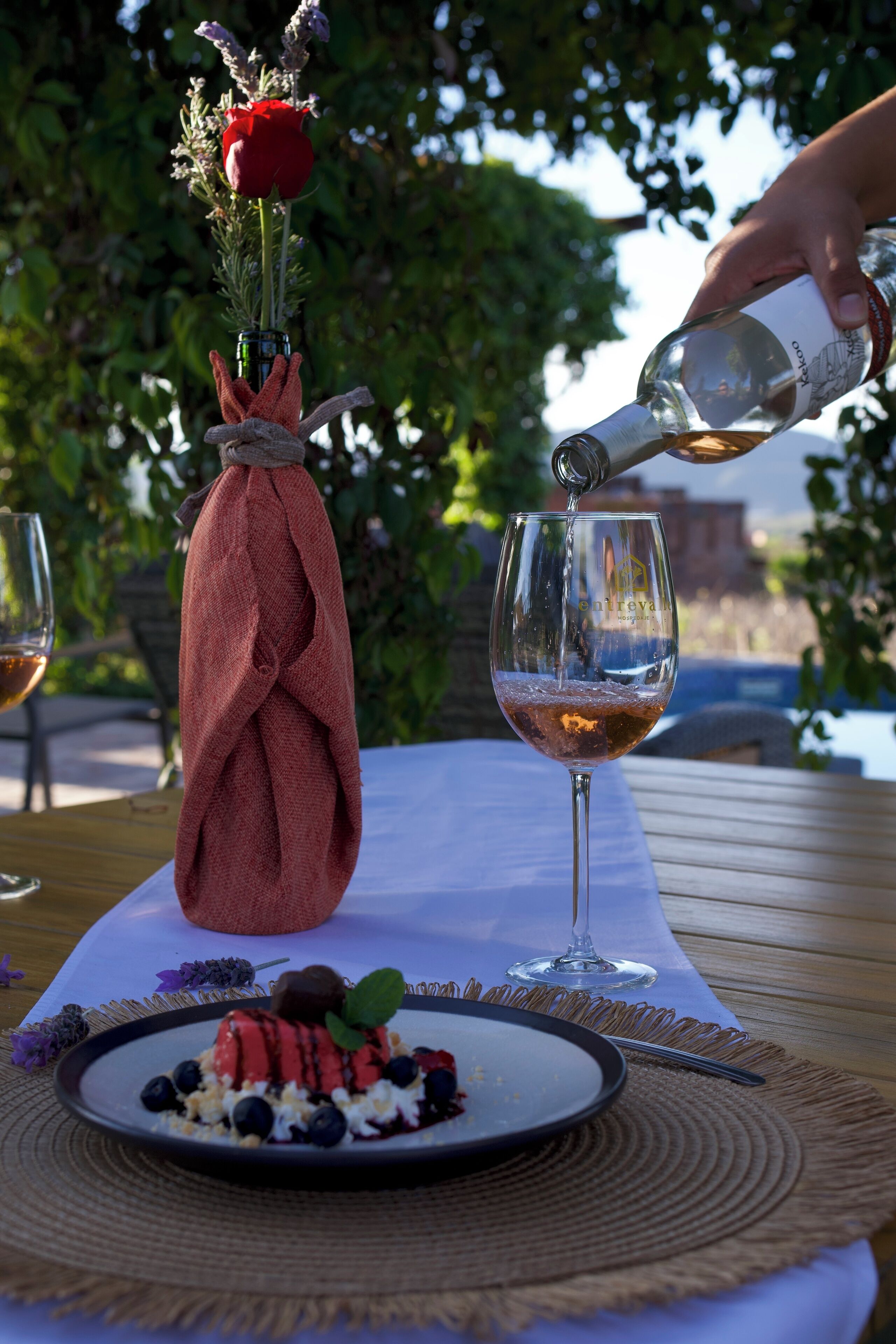 Una mesa con una botella de vino sirviéndose en una copa, un plato con postre y un ramo de flores.