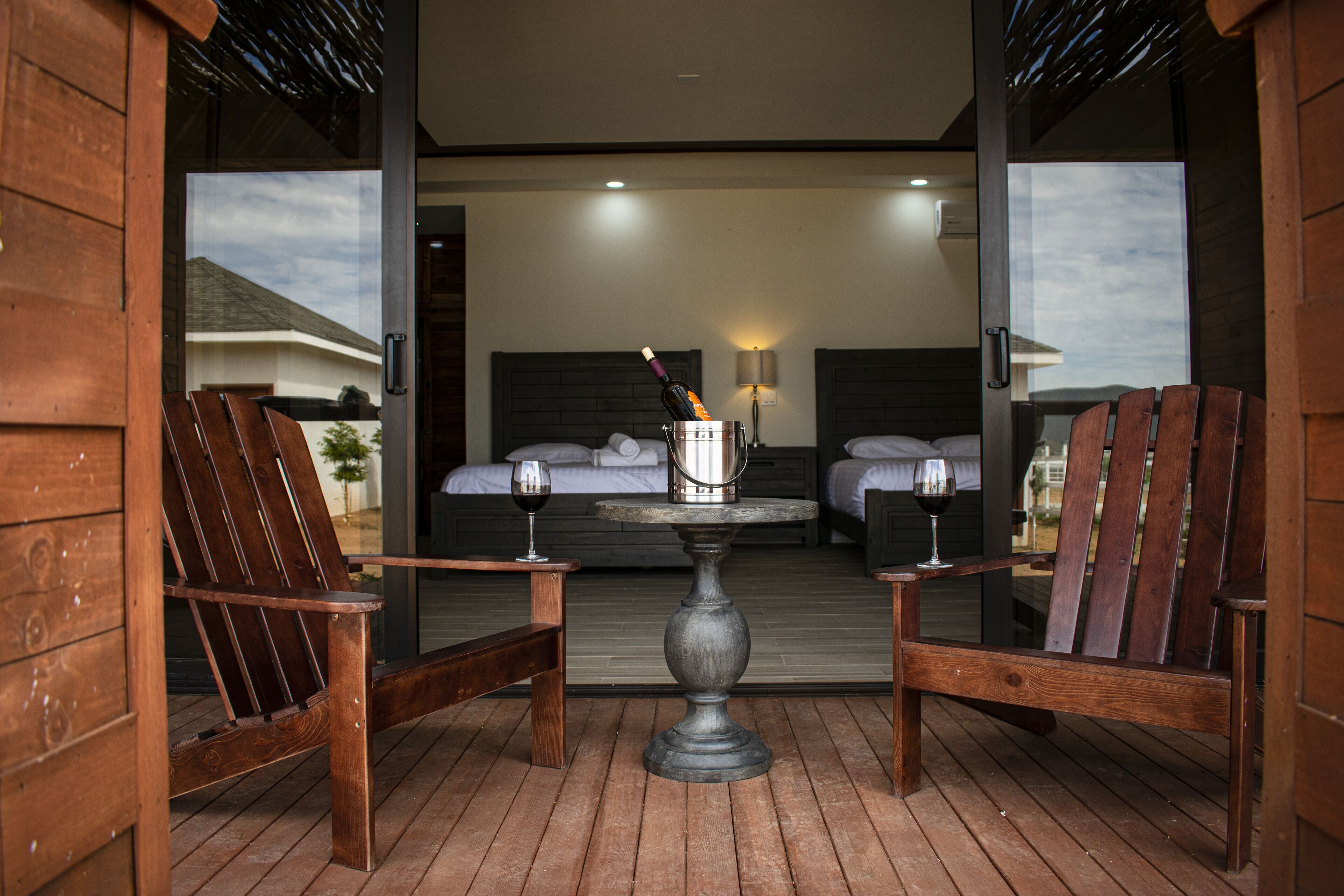 Een houten terras met twee houten stoelen en een tafel, een bed met hoofdeinde en een lamp aan de muur.