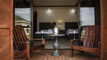 A wooden deck with two wooden chairs and a table, a bed with a headboard, and a lamp on the wall.