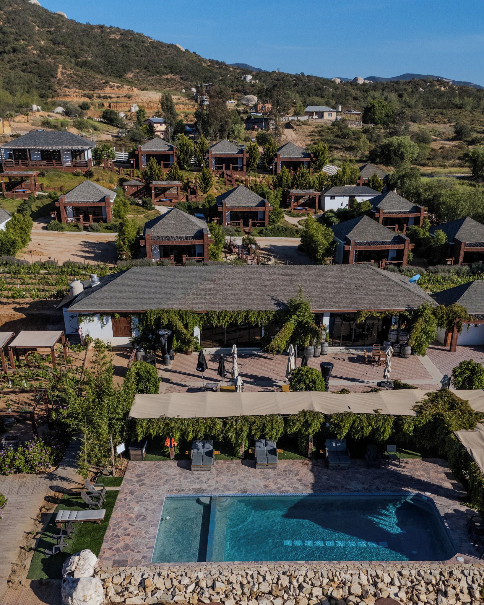 Een resort met meerdere gebouwen in bungalowstijl, een centraal gelegen zwembad en een heuvelachtig landschap op de achtergrond.
