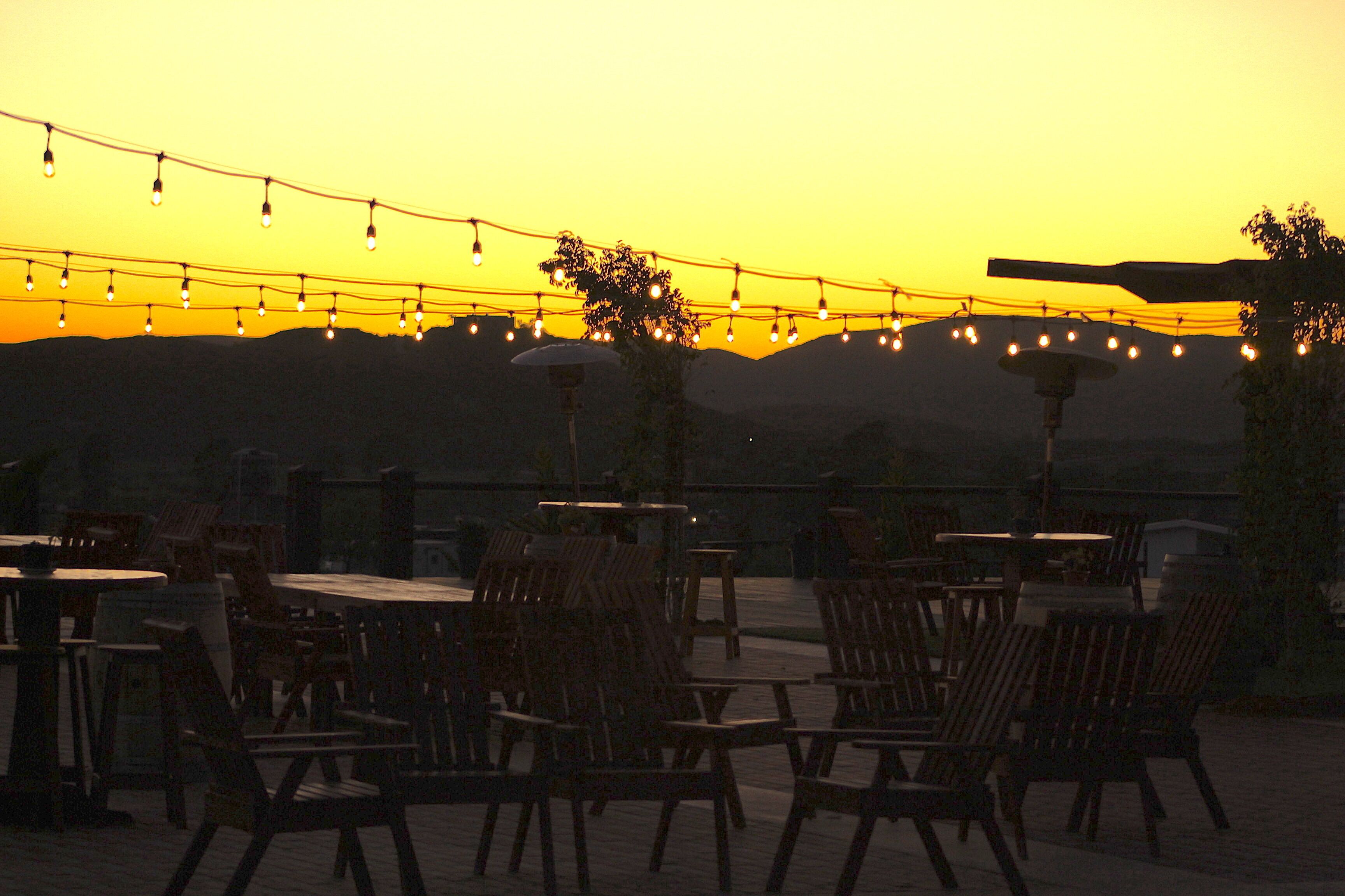 An outdoor seating area with tables and chairs, string lights, and a view of distant hills.