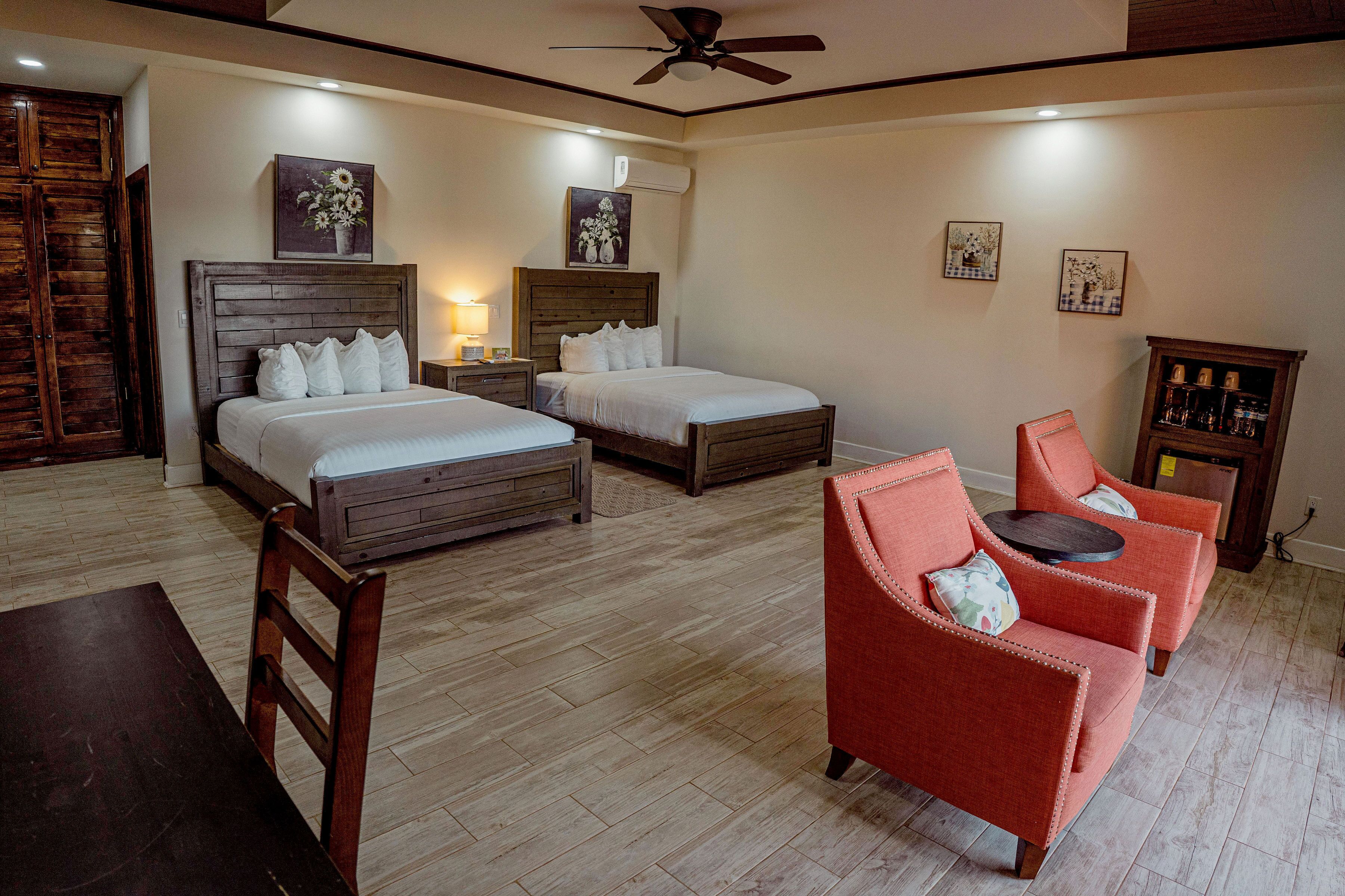 A hotel room with two beds, a wooden headboard, a red armchair, a small table, and a ceiling fan.