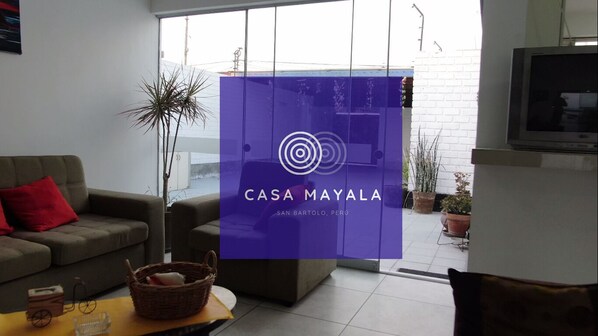 A modern living room with a sofa, a coffee table, and a TV. A purple sign with "CASA MAYALA SAN BARTOLO, PERU" is visible. - Casa Mayala en playa San Bartolo (San Bartolo)