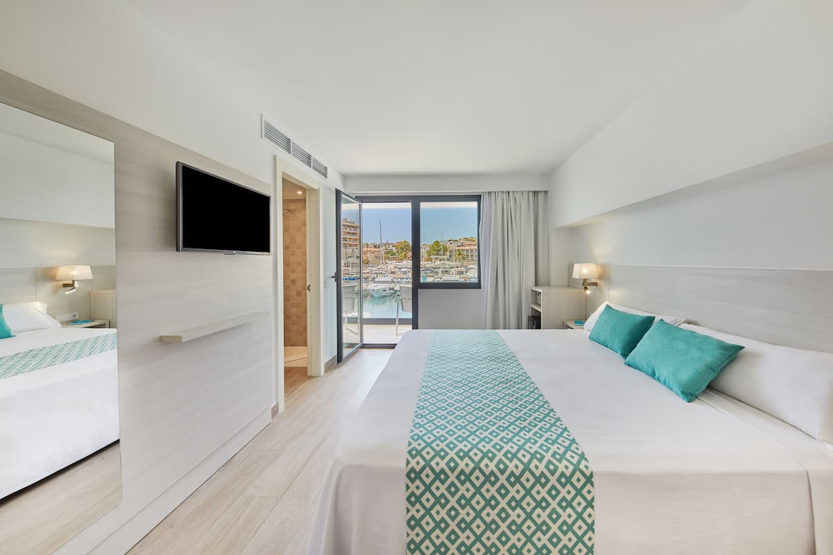 A modern hotel room with a large bed, a TV mounted on the wall, and a balcony with a view of the marina.