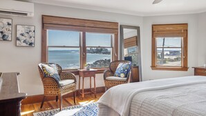 Un dormitorio con una cama grande, dos sillones de mimbre, una mesita y vista al océano a través de amplias ventanas.