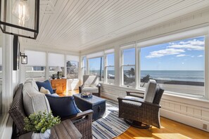 A sunlit room with a view of the ocean, featuring wicker furniture, a blue sofa, and a wooden table. - Ocean/Beach With Magnificent Views:  A Unique New England Experience ! (Kennebunk)