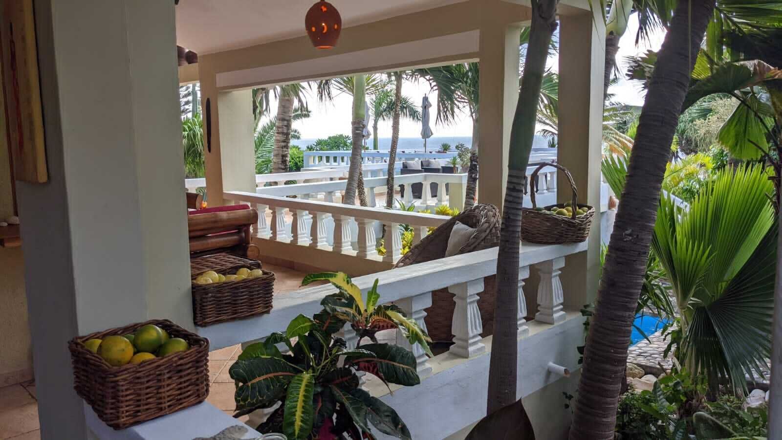 Un balcón con cestos de mimbre, barandal blanco y plantas tropicales.