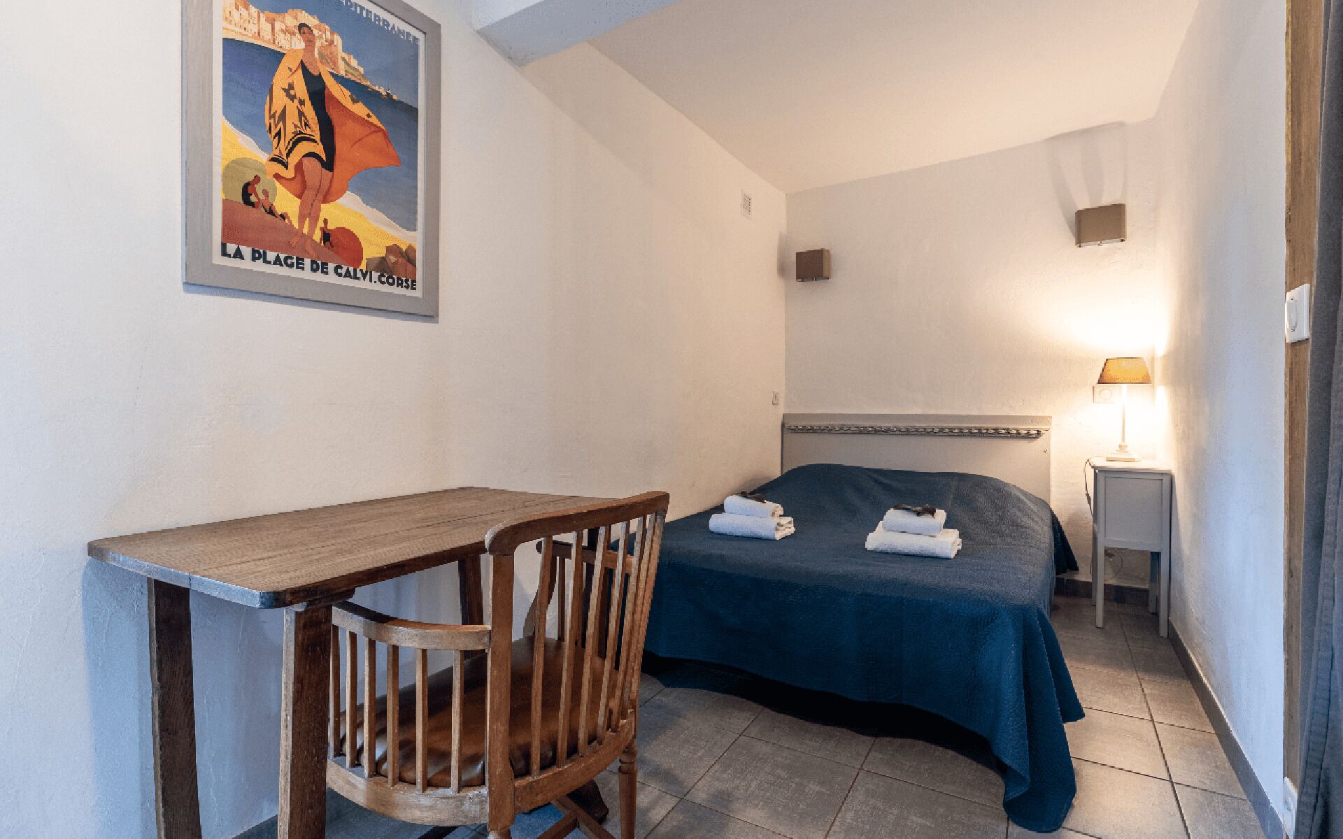 A hotel room with a bed, a desk, a chair, a nightstand, and a lamp.
