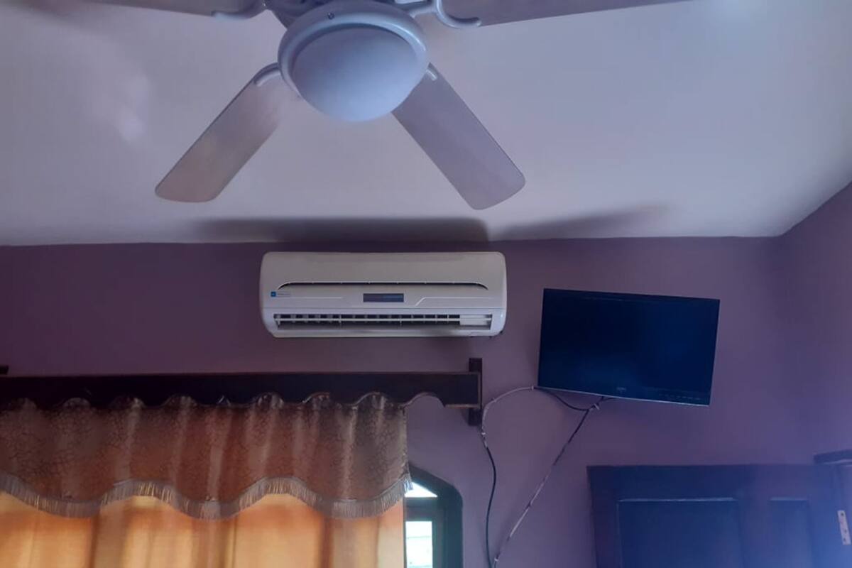 A ceiling fan, an air conditioning unit, a television mounted on the wall, and a window with curtains.