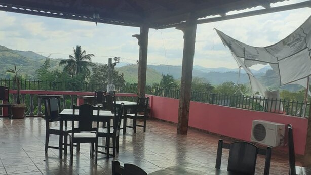 Un patio exterior con suelo de baldosas, mesas y sillas de madera, y vistas a colinas y montañas verdes.