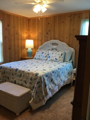 A bedroom with a wooden paneled wall, a bed with a patterned bedspread, a nightstand with a lamp, and a ceiling fan.