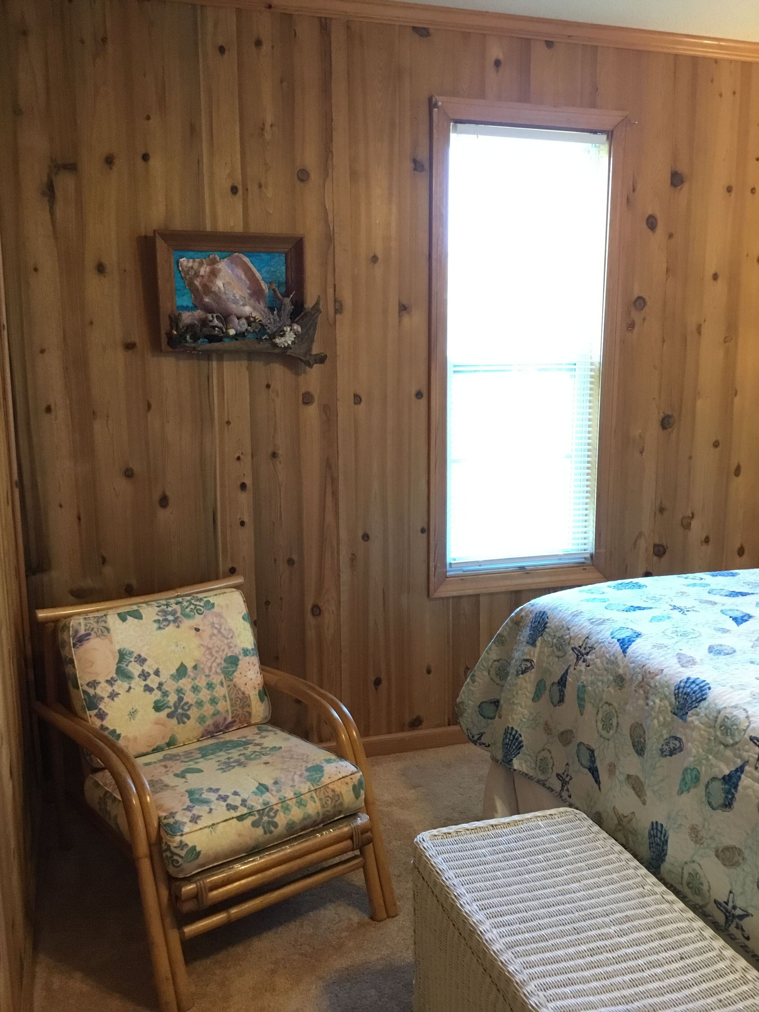 Une pièce aux murs lambrissés, une chaise recouverte d’un tissu à motifs floraux, un lit avec une couvre-lit à fleurs bleues et une fenêtre avec des stores.