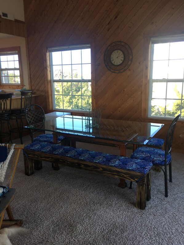 A cozy room with a wooden ceiling, a large window with a view of trees, a round wall clock, and a glass-top table with chairs. - 4 BR Oceanside easy beach access - home feeling! (Salvo)