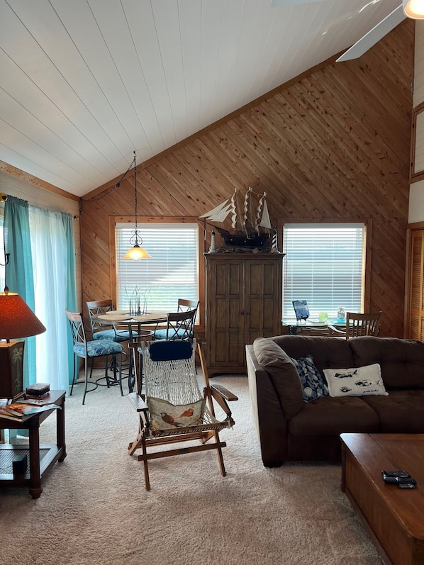 A cozy living room with a high ceiling, wooden walls, a dining area with a table and chairs, and a rocking chair.