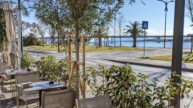 Un café en plein air avec des tables et des chaises, un espace de repos couvert et une vue sur une rivière et un pont.