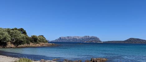 A beach with rocky shoreline, clear blue water, and distant mountains.