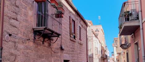 Una estrecha calle adoquinada bordeada de edificios históricos, con balcones adornados con plantas en macetas y un cielo azul despejado.