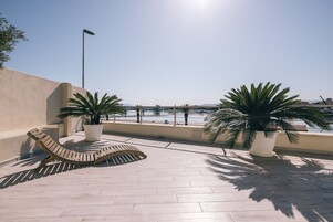 A sunlit outdoor area with a lounge chair, potted plants, and a view of a body of water.