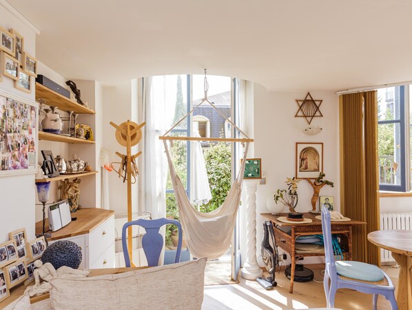 A cozy room with a hammock, a dining table, and a chair. There are shelves with various items and a window with a view of greenery.