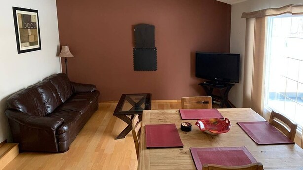 A living room with a brown leather sofa, a wooden dining table with chairs, a TV, and a window with curtains.