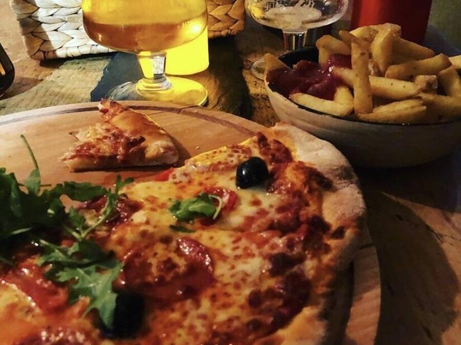 Une pizza garnie, un verre de bière et un bol de frites sur une table en bois.
