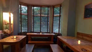 Una habitación acogedora con un banco y una mesa de madera, ventanas amplias con persianas, y un cuadro en la pared.