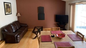 En stue med en brun lædersofa, et træspisebord med stole, et fjernsyn og et vindue med gardiner.