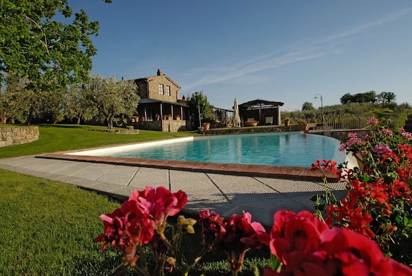 A swimming pool with a stone deck, a stone building with a covered patio, and a grassy area with flowering plants.