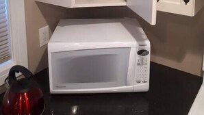 A kitchen with a white microwave on a black countertop, a red kettle, and a cabinet with stacked plates and bowls.