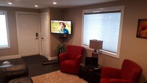 A living room with a large flat-screen TV mounted on the wall, a red armchair, a black leather sofa, and a small side table with a lamp.