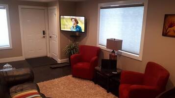 A living room with a large flat-screen TV mounted on the wall, a red armchair, a black leather sofa, and a small side table with a lamp.