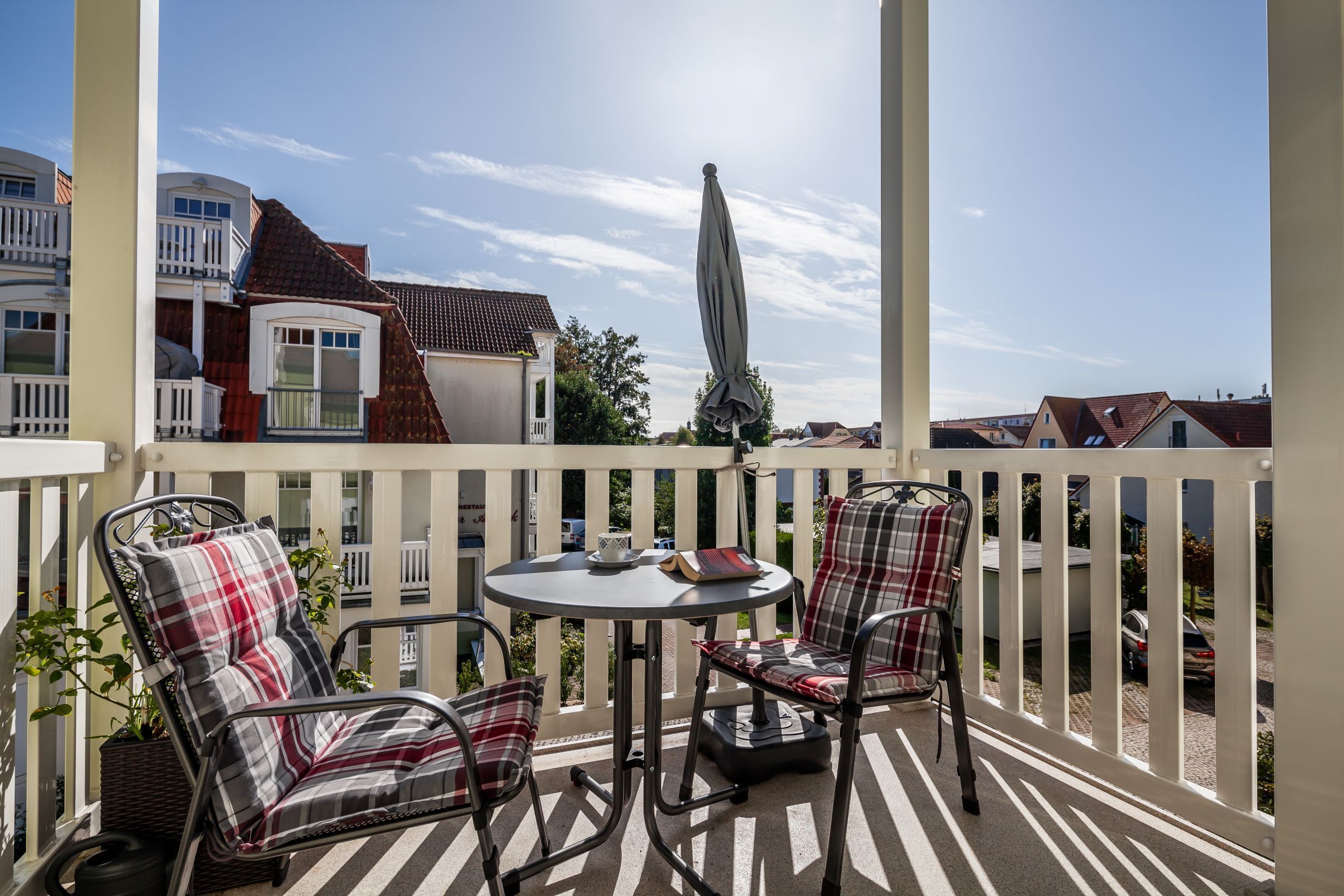 A balcony with two chairs and a table, a white fence, and buildings in the background.