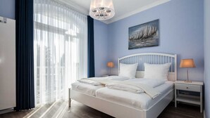 A bedroom with a white bed, two bedside tables, a chandelier, a painting of sailboats, and a window with curtains. - App. Lolita - Lolita (Kühlungsborn)