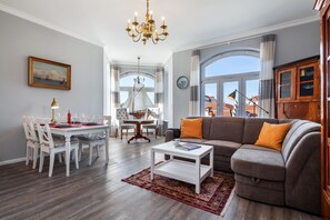 Un salon comprenant un canapé, une table à manger avec des chaises, un lustre et une vue dégagée sur l’extérieur grâce à de grandes fenêtres.