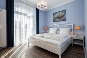 Une chambre à coucher avec un lit blanc, deux tables de chevet avec des lampes, un tableau représentant des voiliers et une fenêtre avec des rideaux légers.